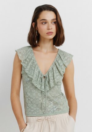 Vrouw met kort bruin haar draagt een lichtgroen kanten topje met gerimpelde halslijn en strik, gecombineerd met beige broek met trekkoord, staande tegen een witte achtergrond.