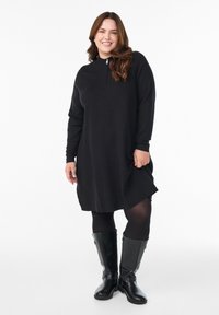 Naeratav naine mustas pikkade varrukatega kleidis, mustades sukkades ja põlve kõrgustes mustades nahkkingades seisab valge tausta vastu.