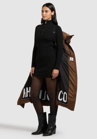 Črna rebrasta obleka z visokim ovratnikom, kombinirana z rjavo, preveliko puhasto jakno z drzno logotipom. Črni gležnjarji zaključujejo videz.