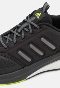 Zapato deportivo negro con cordones grises, parte superior de tela texturizada y tres franjas moldeadas de color gris oscuro. Acentos en verde brillante en la lengüeta y en la suela.