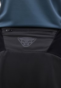 Κοντινό πλάνο μαύρης υφασμάτινης επένδυσης με φερμουάρ και σκούρο γκρίζο λογότυπο προσώπου συμβόλου Puma στο κάτω μέρος της πλάτης, που φοριέται κάτω από ένα μπλε υφασμάτινο πουκάμισο.