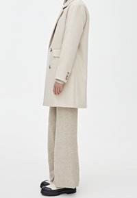 Cappotto beige doppiopetto in tessuto testurizzato, maglione grigio chiaro a maglia sotto, pantaloni crema a gamba larga e scarpe stringate nere e bianche.