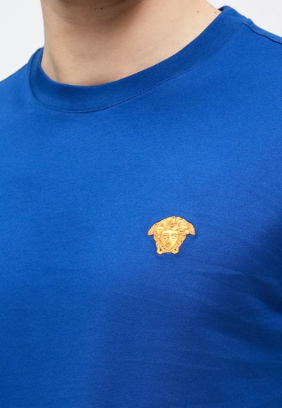 Kék pamut póló környakú kivitelben, bal mellrészen texturált arany hímzett logóval, amely egy klasszikus alakot formáz.