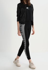 Černá bomberská bunda, přiléhavý černý crop top, legíny s vysokým pasem v černé a šedé barvě s textem "NIKE SPORTSWEAR" a béžové tenisky.