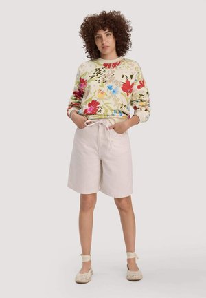 Frau mit lockigem Haar trägt ein langärmeliges Oberteil mit Blumenmuster, beige knielange Shorts mit Bindegürtel und cremefarbene Espadrilles mit Knöchelriemen, steht mit den Händen in den Taschen.