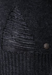 Zwarte gebreide trui met een verouderd patchdetail en een ronde metalen accent. De textuur is geribbeld en combineert gladde en ruwe elementen.