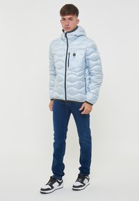 Giacca imbottita azzurro chiaro con design trapuntato, chiusura con zip sul davanti e cappuccio. Presenta dettagli neri e un patch con logo sulla manica. Indossata con jeans.