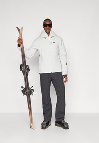 Casaco de esqui branco com capuz, detalhes em preto; calças de esqui cinza escuro; segurando esquis com fixações; usando óculos de sol escuros e botas de esqui.