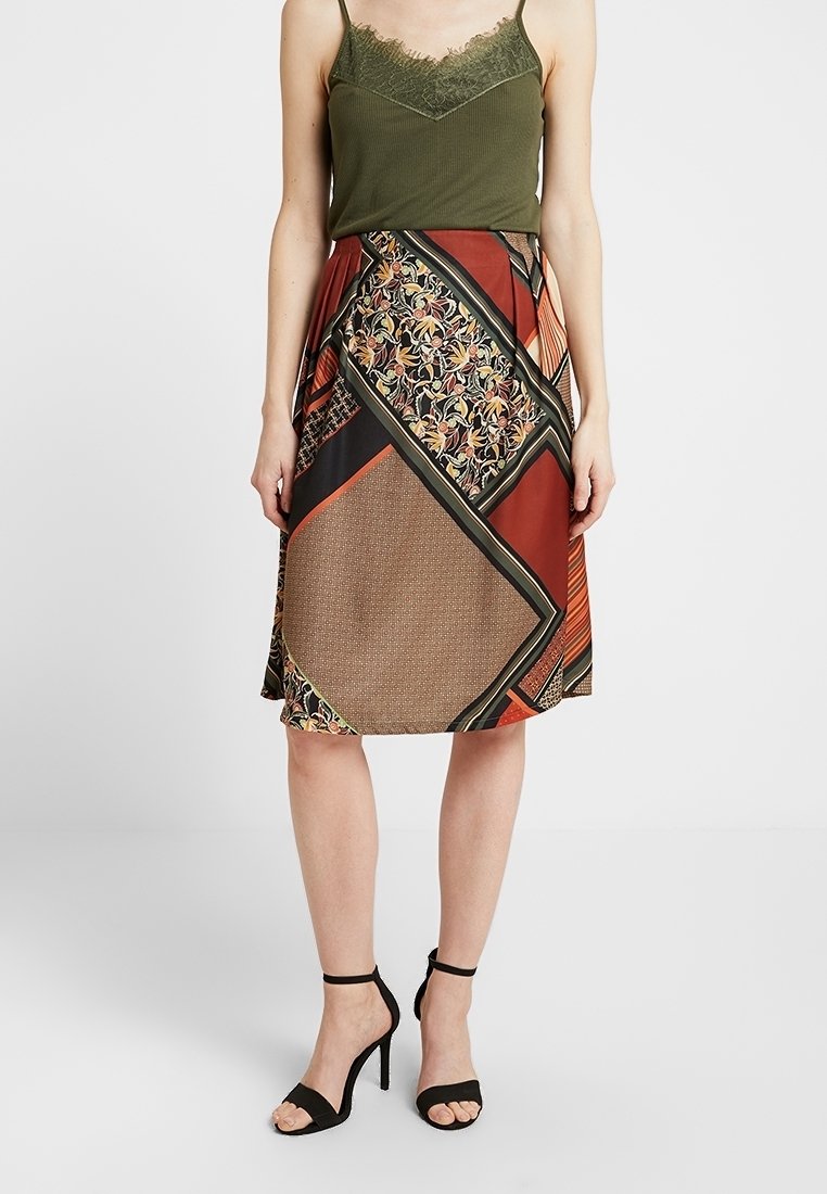 Femme portant un caraco en dentelle vert olive, une jupe patchwork mi-genoux aux tons automnaux, et des sandales noires à lanières à la cheville avec talons hauts.