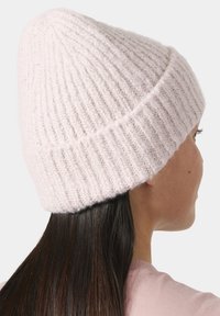 Mujer con cabello largo y oscuro, vistiendo un gorro de punto acanalado rosa claro y una camiseta rosa pálido, de perfil contra un fondo liso.