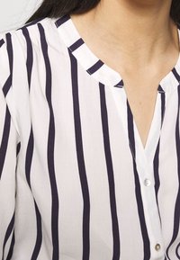 Blouse blanche à rayures verticales bleu marine, fermeture à boutons et col arrondi. Le tissu semble léger et lisse avec une légère brillance.