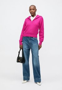 Rózsaszín kötött pulóver, cipzáras nyakkal, kék bő szabású farmernadrággal párosítva. A fekete kézi táska és a fehér hegyes orrú cipők teszik teljessé az öltözéket.