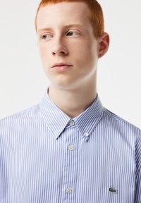 Camisa de botones a rayas azul y blanca con cuello, que presenta un pequeño logotipo verde bordado en el lado izquierdo del pecho y botones blancos.