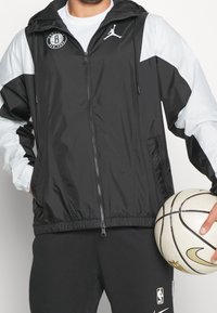 Sort og lys grå vindjakke med lynlås, hætte og elastiske manchetter, der har et logo på brystet. Holder en basketball.