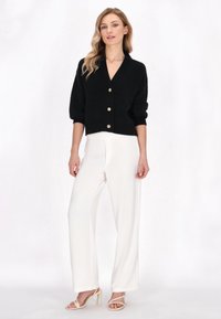 Cardigan corto nero con scollatura a V e bottoni frontali, abbinato a pantaloni larghi bianchi. Modella con tacchi a strappo beige.