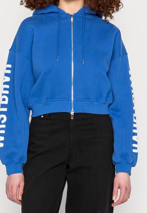 Persona que lleva una sudadera con capucha azul con cremallera y texto blanco en las mangas y pantalones negros, de pie contra un fondo liso.