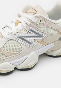Beige and white sneaker with mesh and suede panels, featuring a prominent silver logo, flat laces, and a textured rubber sole.