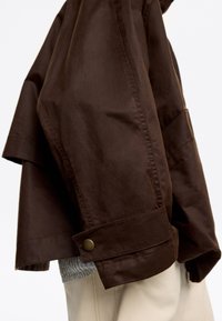 Primer plano de una persona que lleva una chaqueta marrón oscura sobre un suéter gris y pantalones beige claros, mostrando detalles de costura y un botón de latón en el puño.