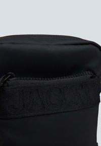 Sort stofpose med en glat tekstur og en lynlåslukning. Øverste del har et struktureret bånd med "JACKIT" præget. Kompakt cylindrisk form.