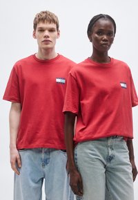 T-shirts vermelhas de algodão com mangas curtas, apresentando um emblema azul e branco no peito esquerdo. Usadas com jeans de ganga azul claro.
