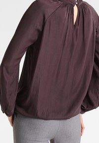 Mørk lilla bluse med nøglehul i ryggen, samlet halsudskæring og lange, løse ærmer. Fremstillet af let, flydende stof.