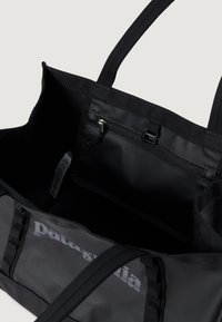Geantă tote din nylon negru cu curele lungi, dotată cu un buzunar interior cu fermoar și logo-ul "Patagonia" în gri pe o parte. Textură netedă, mată.