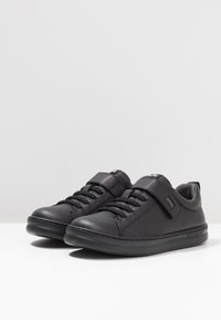 Mustad sünteetilised tossud mustas värvuses, millel on tasane tald, paelte ja Velcro rihmadega sulgurid, sile tekstuur ja minimalistik disain.