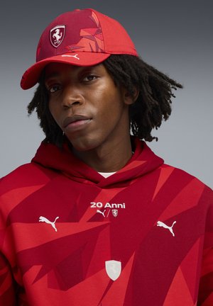 Rote Kapuzenjacke mit geometrischen Mustern, weißen Akzenten und Logo-Details. Die passende Mütze ist rot mit einem stilisierten Logo und kantigem Design.