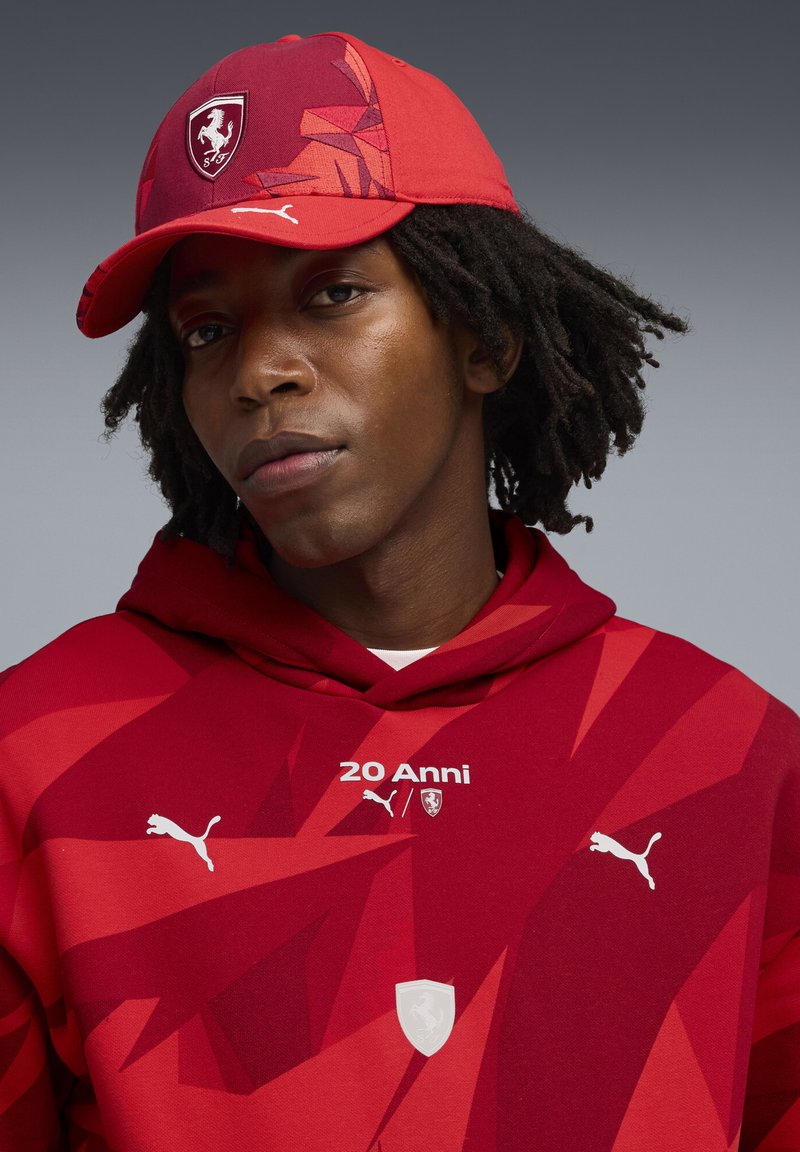 Czerwona bluza z kapturem z geometrycznymi wzorami, białymi akcentami i detalami logo. Pasująca czapka jest czerwona z stylizowanym logo i kątowym wzornictwem.