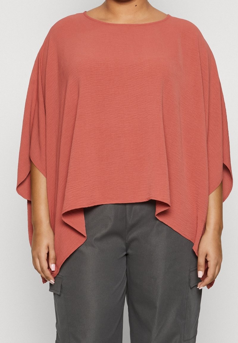 Haut surdimensionné couleur rouille avec un col rond et un design fluide, confectionné dans un tissu texturé. Porté avec un pantalon cargo gris.