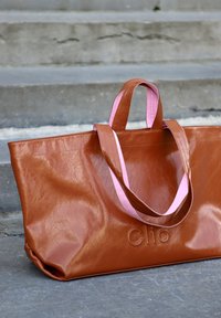 Sac fourre-tout en cuir synthétique marron avec des poignées roses. Présente un logo en relief "Clio" sur le devant. Texture lisse et design spacieux.