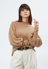 Beige gerippter Pullover mit Ballonärmeln, der Knotendetails und einen hohen Kragen hat. Kombiniert mit cremigen Hosen und einem schwarzen Gürtel mit goldenen Schnallen.