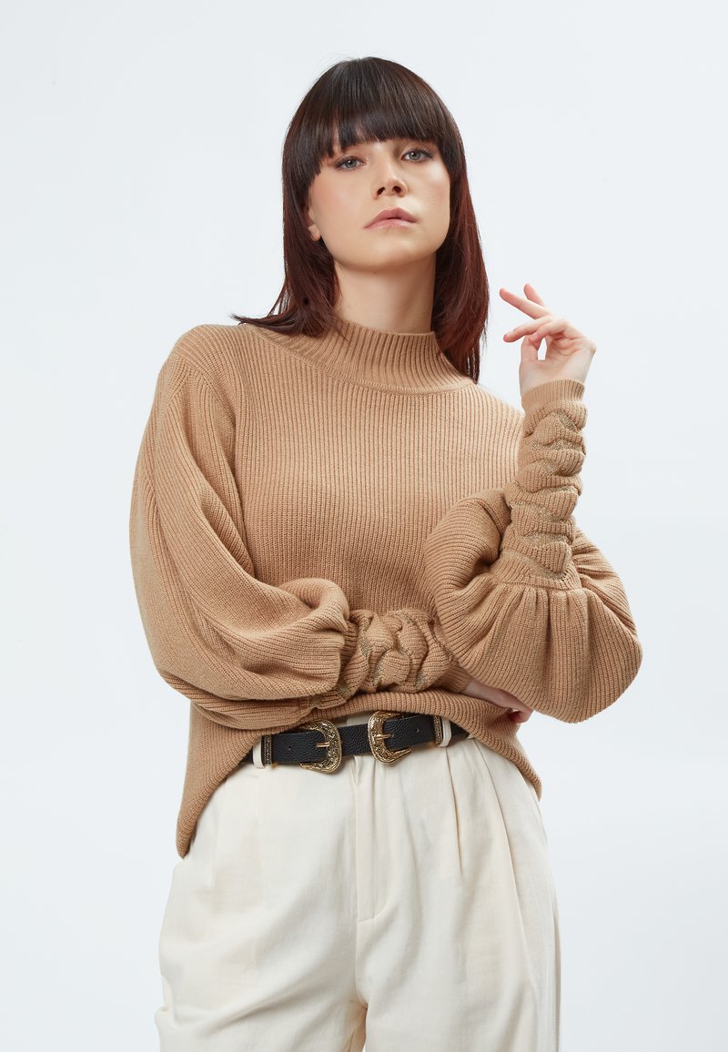 Beige gerippter Pullover mit Ballonärmeln, der Knotendetails und einen hohen Kragen hat. Kombiniert mit cremigen Hosen und einem schwarzen Gürtel mit goldenen Schnallen.