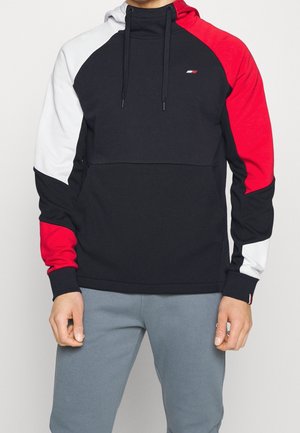 Mann trägt einen Color-Block-Hoodie mit schwarzen, roten und weißen Paneelen, kombiniert mit hellgrauen Hosen, steht vor einem einfarbigen Hintergrund.