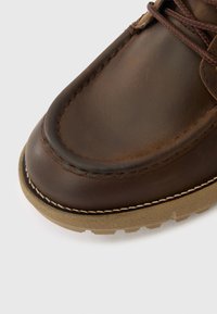 Brūna ādas kurpe ar noapaļotu purngalu, izceļas ar kontrastējošām šuvēm un fakturētu gumijas zolīti. Šņorītes ir sasietas virs mēlītes.