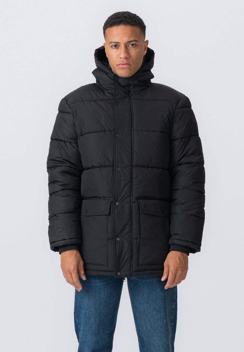 Sort pufferjakke med høj krave og hætte, med knaplukning, to frontlommer og quiltet design. Båret over blå jeans.