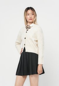 Cárdigan de punto de cable crema con botones negros, de largo corto, con puños acanalados, usado sobre una camisa de cuello a rayas, combinado con una falda negra plisada.