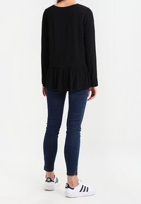 Maglietta nera a maniche lunghe con orlo a peplo, indossata con jeans skinny blu scuro e scarpe da ginnastica bianche e nere, vista da dietro.