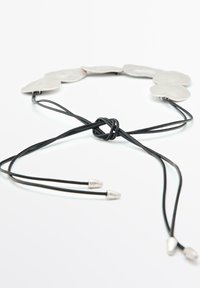 Collana in metallo argento con forme di piastra irregolari e lucide. Corda nera regolabile con punte metalliche argentate, fissata con un nodo.