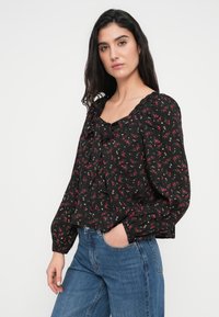 Μαύρο φλοράλ μπλουζάκι με λαιμόκοψη με βολάν, μακριά φουσκωτά μανίκια και φιόγκο στο μπροστινό μέρος. Φέρει μικρά ροζ και λευκά λουλούδια.