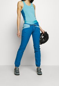 Conjunto deportivo azul que incluye una camiseta sin mangas y pantalones azules contrastantes con cordón. El modelo sostiene un casco negro.