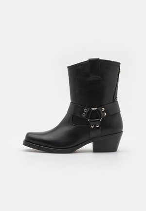 Botte de cowboy en cuir noir montant jusqu'au milieu du mollet avec bride à harnais et clous en argent, talon empilé et languette de traction sur fond blanc.