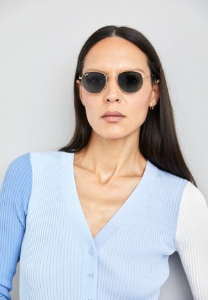 Ochelari de soare rotunzi cu lentile întunecate și rame aurii. Modelul poartă un cardigan ribbed de culoare albastru deschis cu nasturi și mâneci albe în contrast.