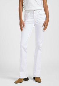 Jeans de mezclilla blanca de pierna acampanada con lazos para cinturón, cinco bolsillos y cierre de botón, combinados con botas de tacón en tono beige. Textura suave y corte ajustado.
