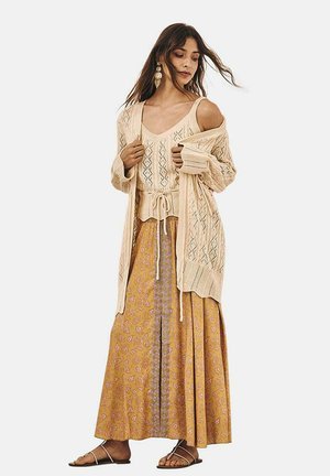 Cárdigan de punto beige sobre una blusa beige con estampado y una falda maxi amarilla con flores. Presenta una cintura con cordón y tejidos texturizados. Sandalias planas.