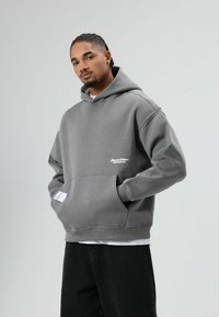 Grijze hoodie van zachte stof met een kangoeroezak, geribbelde manchetten en een klein logo op de borst. Draagt over een wit shirt en gecombineerd met zwarte broek.