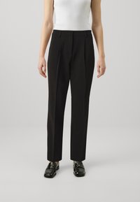 Pantalon noir sur mesure à taille haute, avec une coupe droite et des plis subtils, associé à des mocassins noirs brillants.