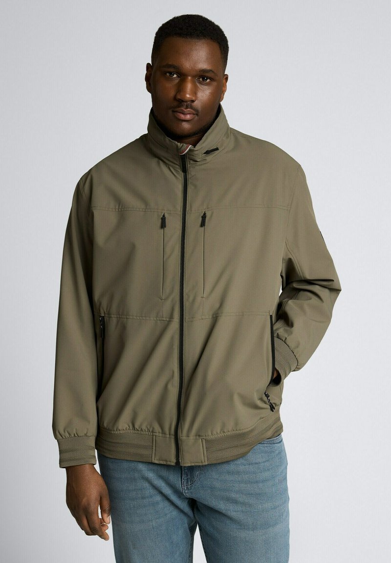 Mężczyzna w oliwkowozielonej kurtce na zamek z wysokim kołnierzem i jasnoniebieskich dżinsach, stojący na tle jednolitego szarego tła.