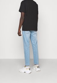 Schwarzes Baumwoll-T-Shirt, hellblaue Jeans und weiße Sneakers mit goldenen Akzenten, von hinten vor einfarbigem Hintergrund gesehen.