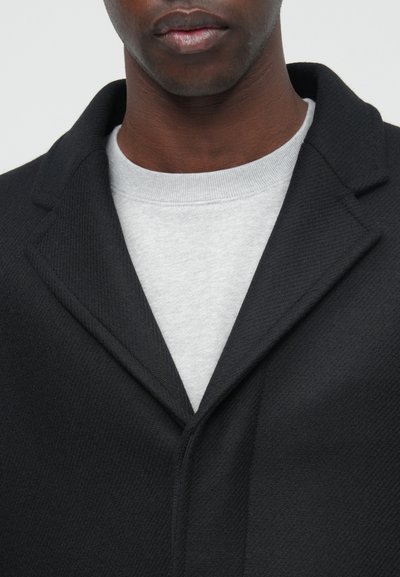Fekete kabát texturált felülettel és kihajtott gallérral, egy világosszürke, kerek nyakú pulóver felett viselve.